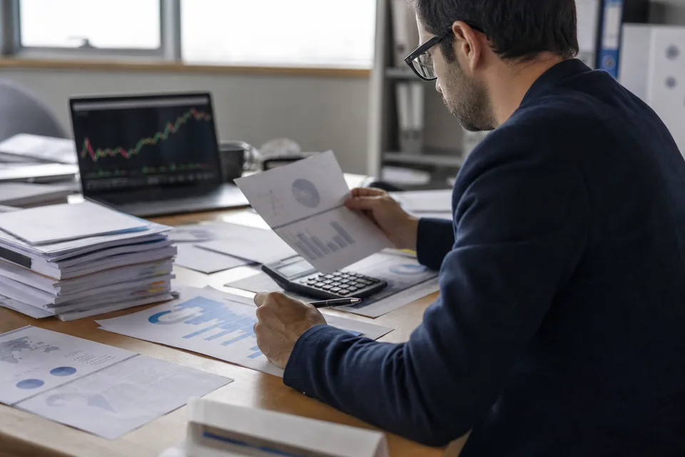 Financial analyst desk with printed balance sheets, a calculator and stock charts on a monitor